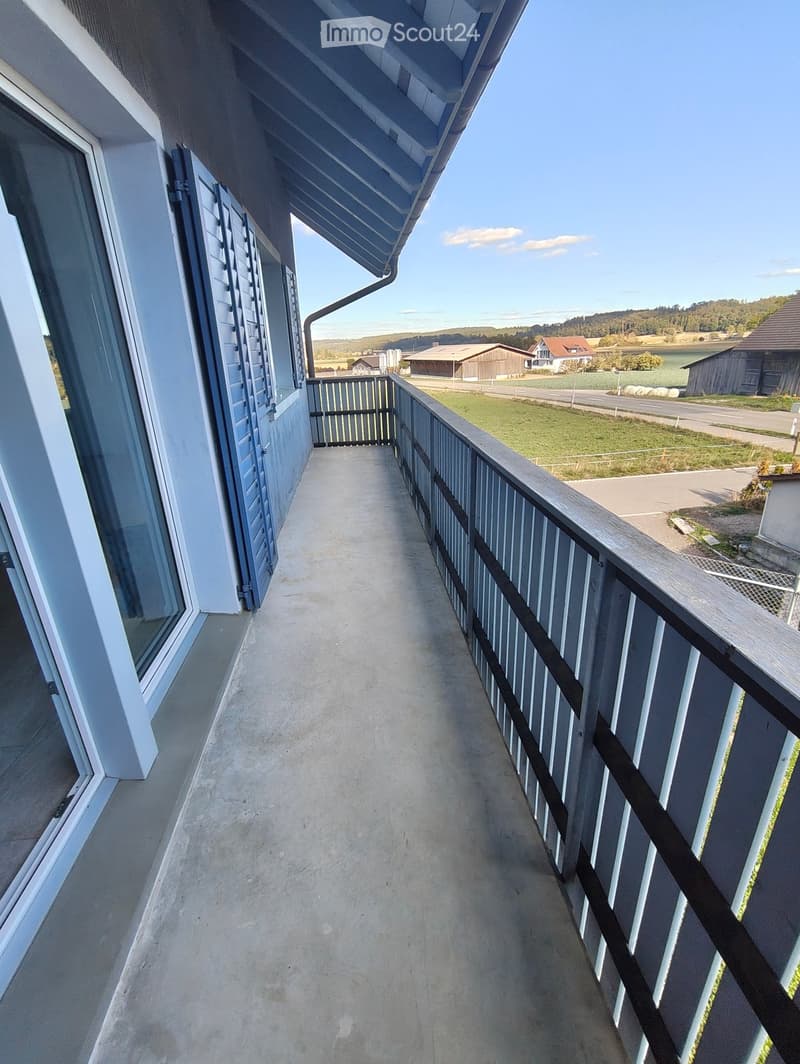 Balcony with wooden railing, overlooking a rural landscape with fields, houses, and a body of water.