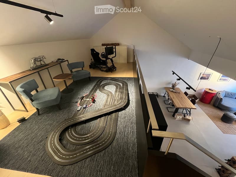 A multi-level room with a toy car track in the foreground, a gaming chair, a sofa, and a small table with a lamp. The room features modern decor and ample natural light.