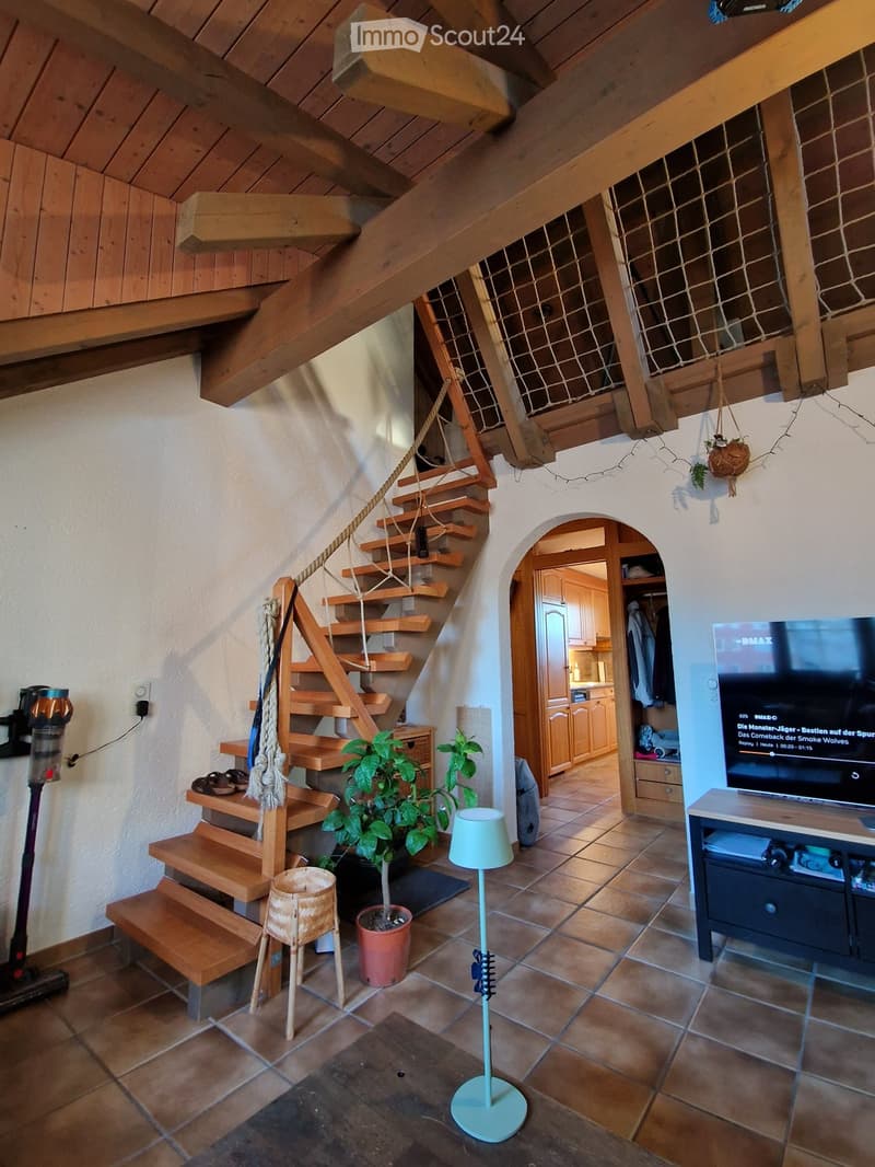 A cozy living area with a wooden staircase leading to an upper level, a television on a stand, a small table with a lamp, and potted plants.