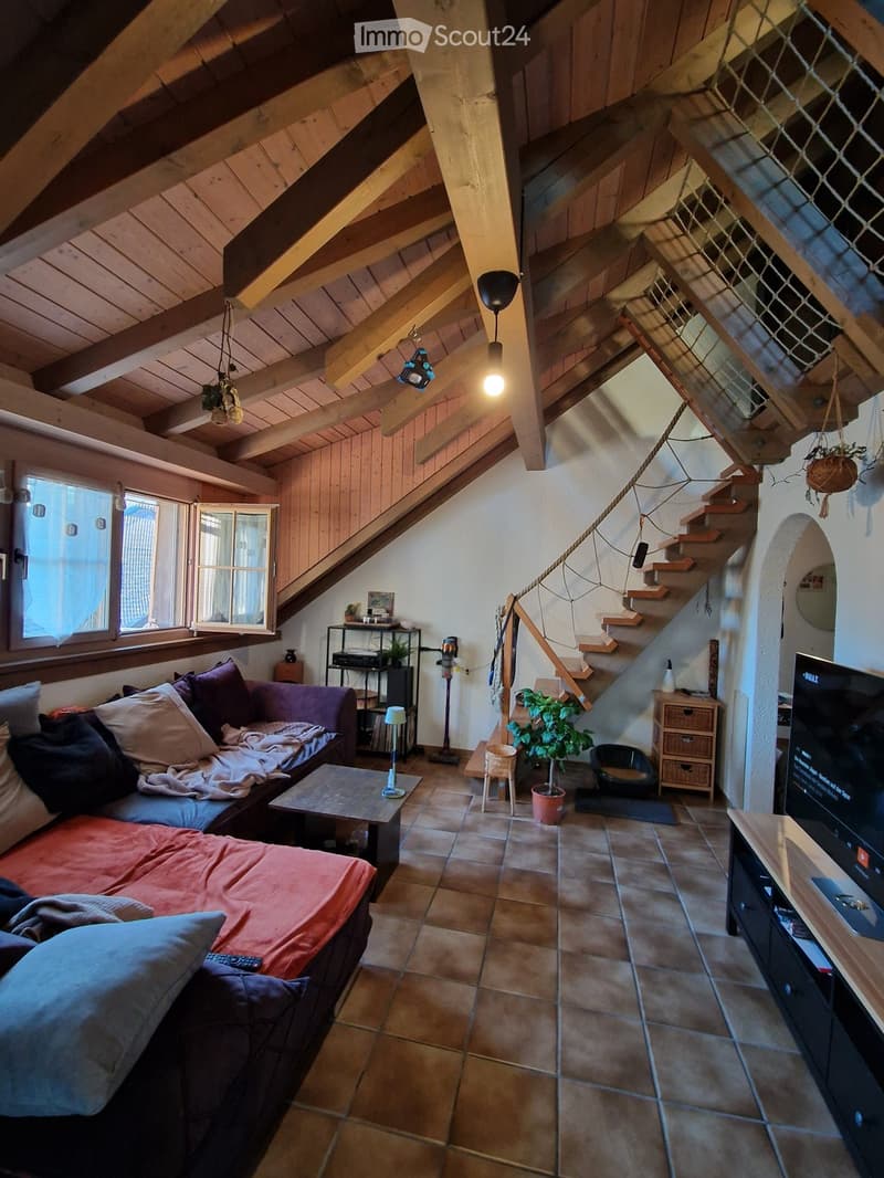 Cozy living room with exposed wooden beams, a large window, a staircase leading upwards, a sofa with pillows, a coffee table, a television, a small table lamp, a potted plant, and a cat bowl.