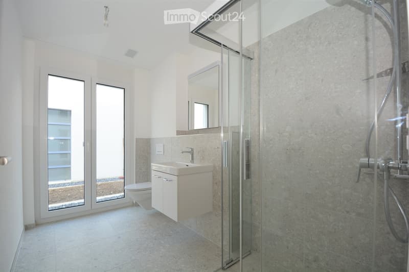 Modern bathroom with a white sink, toilet, mirror, and a glass-enclosed shower. The room features large windows and light-colored tiles.