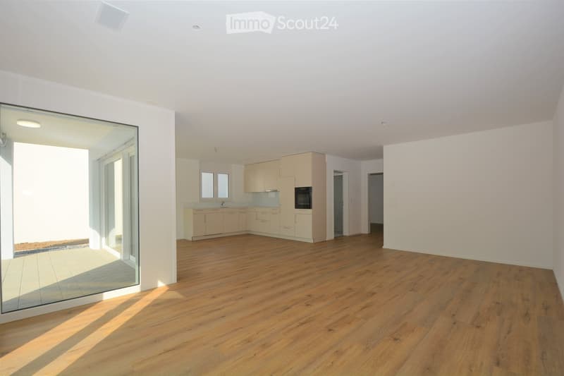 Bright, open-plan living and dining area with wooden flooring, large glass sliding doors leading to a balcony, and a modern kitchen with light-colored cabinets and stainless steel appliances.