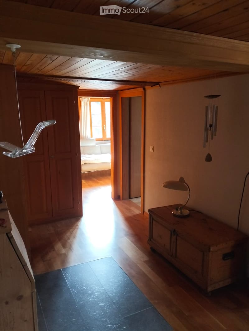 A cozy, wooden-floored corridor with a small table and lamp, leading to a bedroom with a sofa and window view.