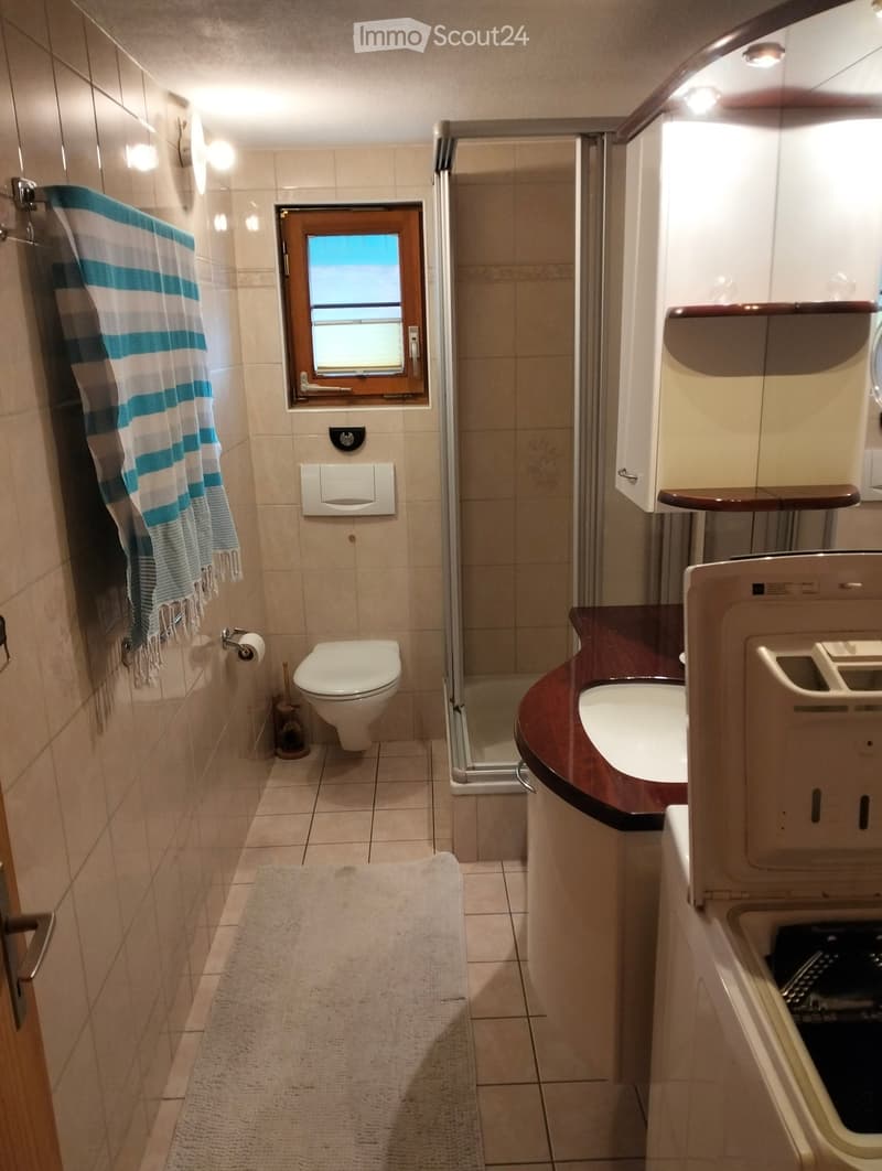 Modern bathroom with beige tiles, a toilet, a sink with a dark countertop, a shower area with a glass door, a towel on the wall, and a small window above the toilet.
