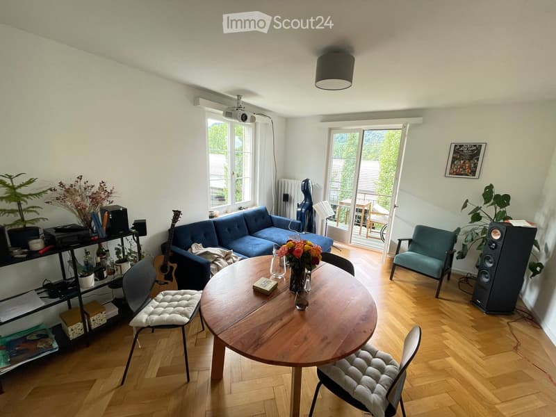 A modern living room with wooden flooring, a blue sofa, a round wooden table, chairs, a guitar, speakers, and plants. There is a large window and a sliding glass door leading to a balcony.