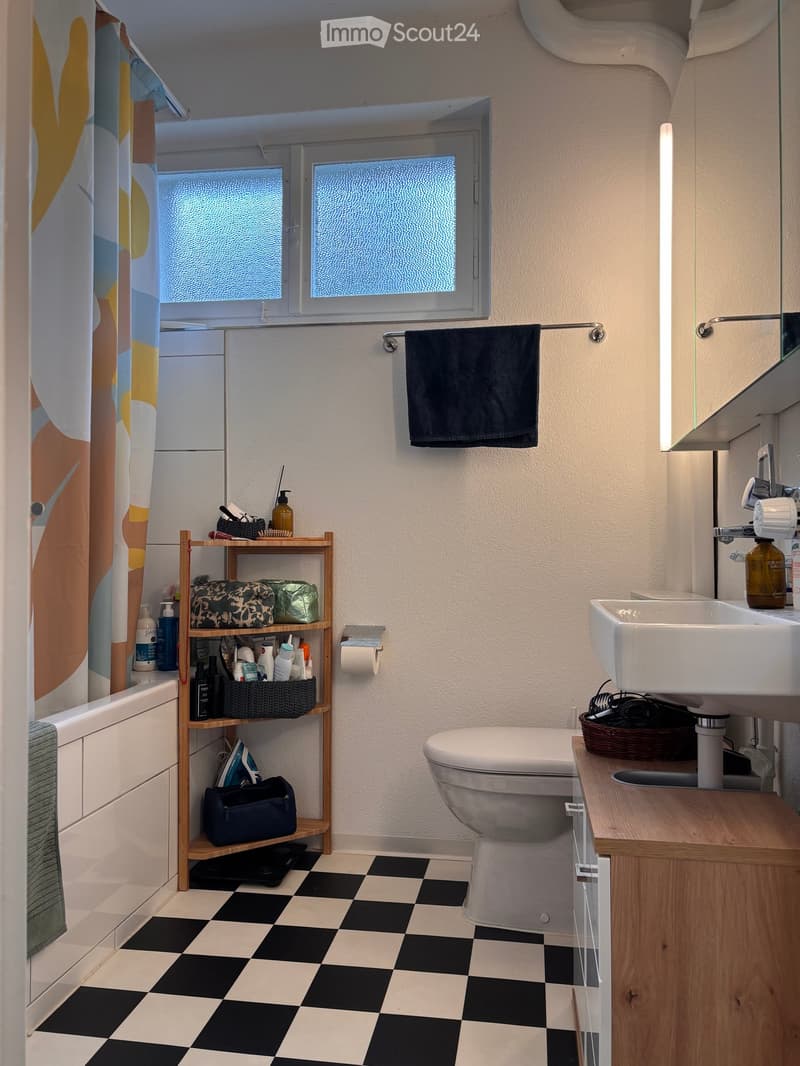 Bathroom with black and white checkered floor, white toilet, sink, bathtub, wooden shelf with various items, towel, and mirror.