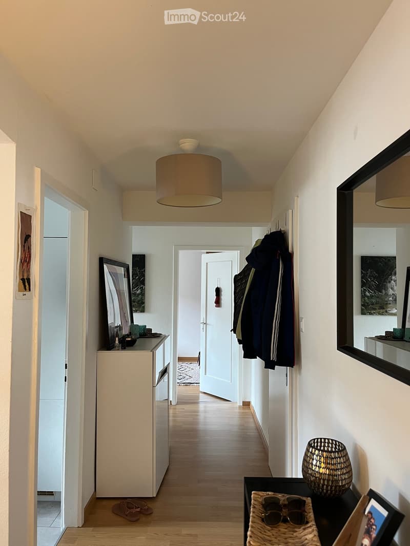 A modern hallway with light wooden flooring, white walls, and a ceiling light fixture. There are several doors leading to other rooms, a coat rack with jackets, and a small table with a decorative bowl and framed pictures.