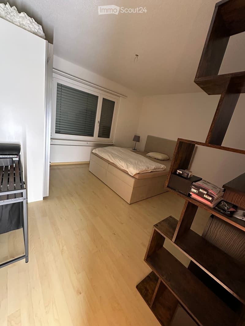 A modern bedroom with a wooden floor, a bed with white bedding, a nightstand with a lamp, and a wooden shelf with books and other items.