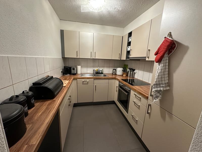 Modern kitchen with light-colored cabinets, wooden countertops, and various appliances including an oven, sink, coffee maker, and toaster. A red oven mitt and a towel are hanging on the wall.