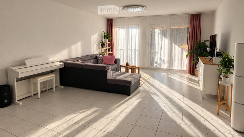 Bright living room with a grey sectional sofa, white piano, small wooden table, and a TV stand with a screen. Natural light streams in through large windows with sheer curtains.