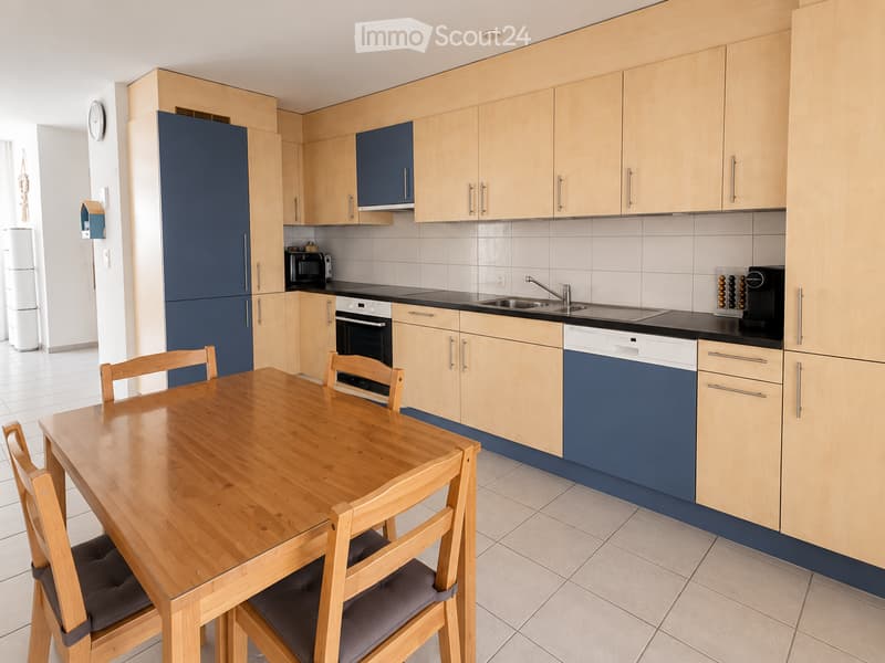 Modern kitchen with light wood cabinets, dark blue lower units, a wooden dining table with four chairs, and various appliances including a refrigerator, oven, dishwasher, microwave, and coffee maker.