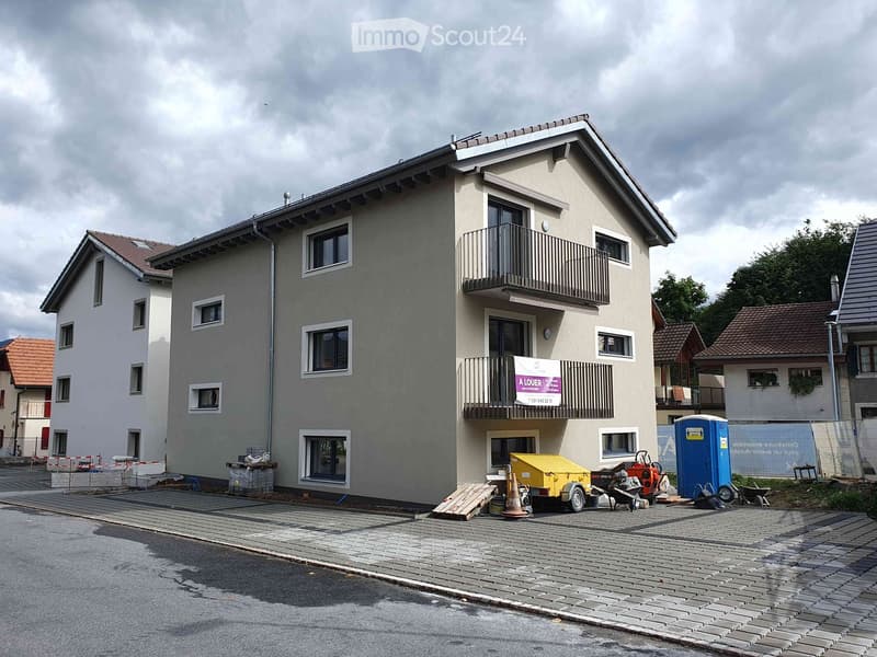 Two-story house with grey facade, multiple windows, and a balcony on the upper floor. The building is surrounded by a paved area with construction materials and equipment.