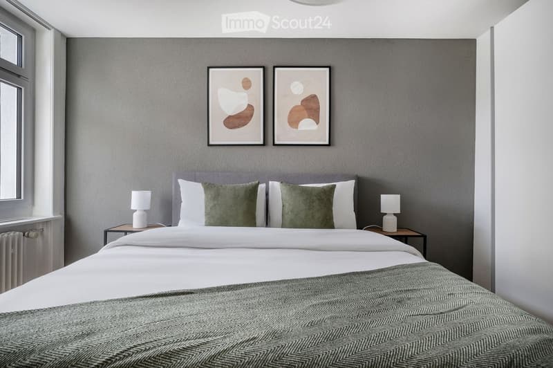 Modern bedroom with a large double bed, white linens, green throw pillows, two nightstands with lamps, and two abstract art pieces on the wall.