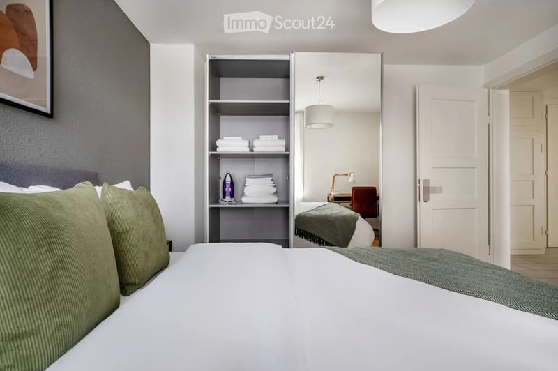 Modern bedroom with a neatly made bed, green pillows, a grey blanket, a built-in wardrobe with folded towels and an iron, a small desk with a chair, and a hanging lamp.