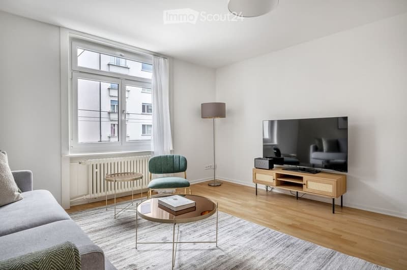 Modern living room with a grey sofa, coffee table, armchair, TV, floor lamp, and a small round table with a book on it.