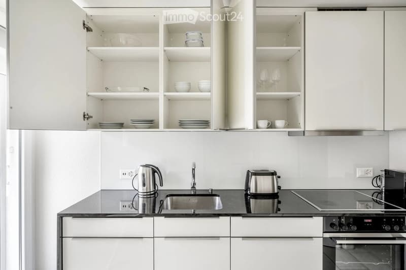Modern kitchen with white cabinets, stainless steel appliances, and a black countertop. Visible items include a sink, electric kettle, toaster, and various dishes in the cabinets.