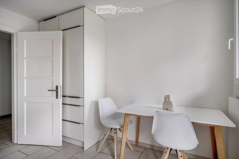 A modern, minimalist room with a white table, two white chairs, and a decorative vase on the table. The room features white walls, a white door, and a tiled floor.