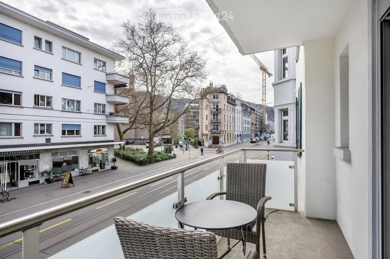 Balcony with a round table and two wicker chairs, overlooking a street with buildings and a tree-lined sidewalk.