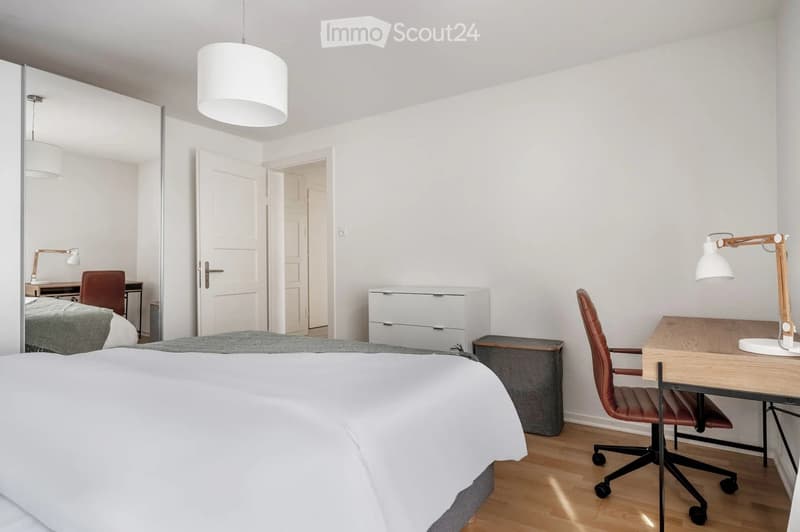 Modern bedroom with a large bed, a desk with a chair, a mirror, and a hanging pendant light. The room features light-colored walls and wooden flooring.