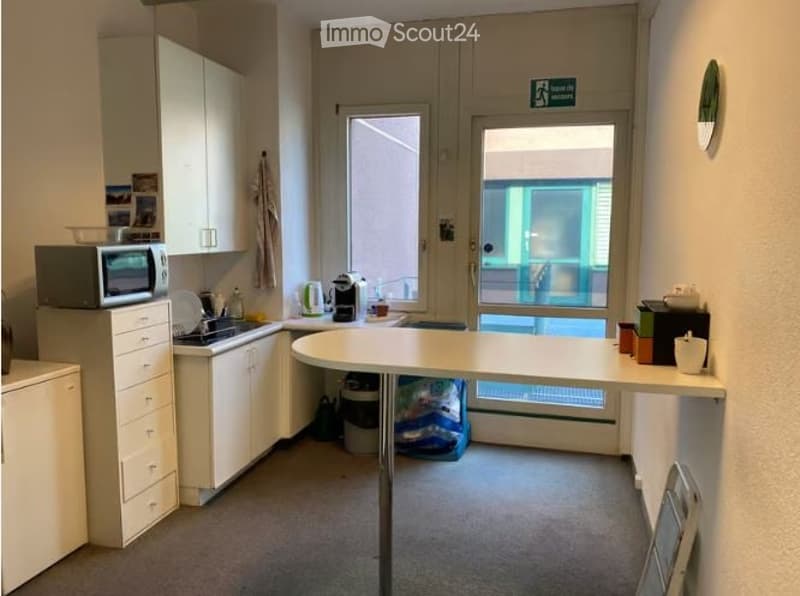 A small, modern kitchen area with white cabinets, a microwave, a sink, and a small table. There is a window and a door leading outside.