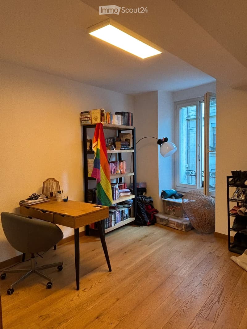 A cozy, well-lit room with wooden flooring, a desk with a chair, a bookshelf filled with books and a colorful flag, a wicker ball, and a shoe rack near the window.