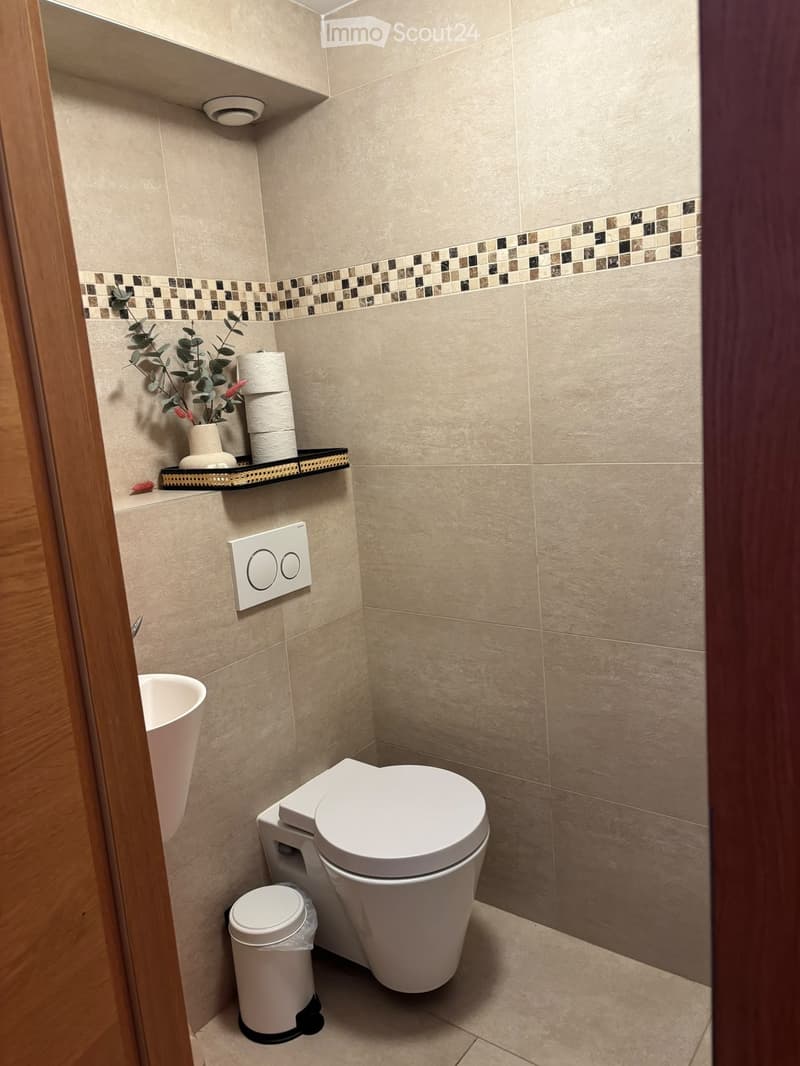 Modern bathroom with beige tiles, a toilet, a sink, a small trash can, and a decorative shelf holding toilet paper and a plant.