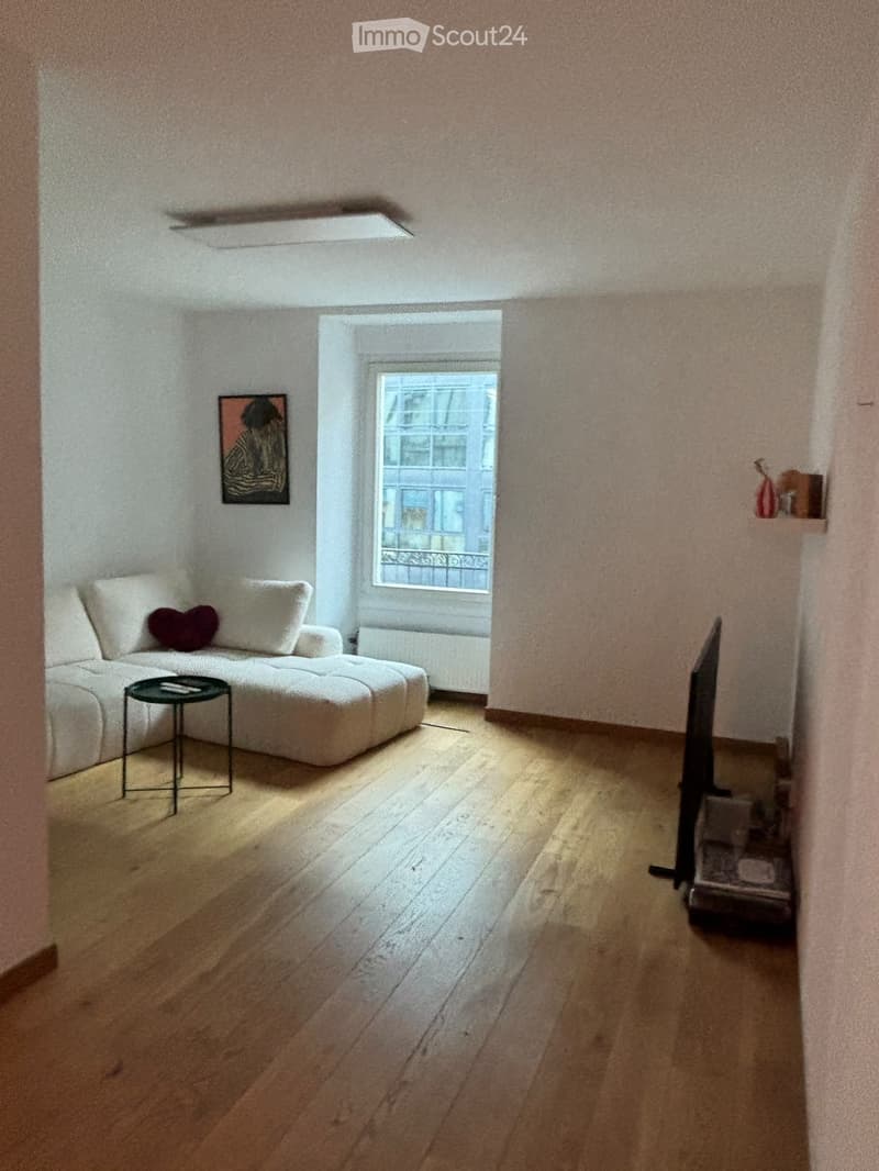 Living room with light wooden flooring, white walls, a large window, a white sectional sofa with a heart-shaped pillow, a small black coffee table, and a picture frame on the wall.