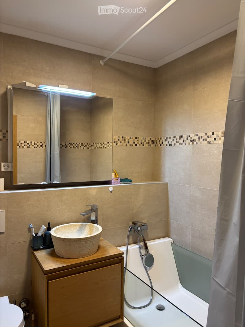 Modern bathroom with a wooden vanity, a round sink, a bathtub with a handheld shower, and a large mirror with built-in lighting.