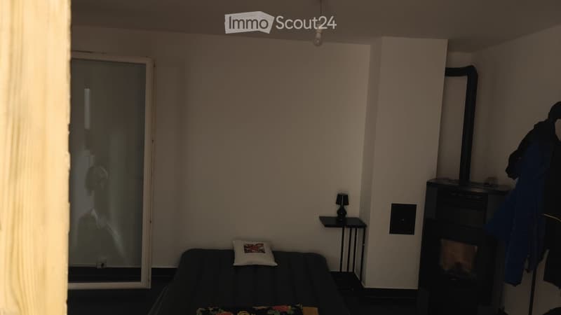 A small, modern living room with a dark-colored sofa, a decorative pillow, a small side table with a lamp, and a coat rack with jackets.
