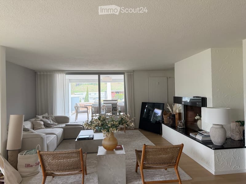 Modern living room with a grey sectional sofa, coffee table, wicker chairs, a large TV, and a fireplace with decorative items. Large windows offer a view of the outdoor area.