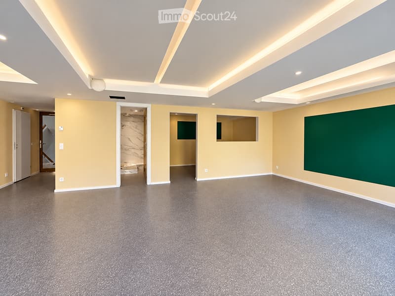 Empty room with light yellow walls, grey carpeted floor, and white ceiling with recessed lighting. Features several doors and windows leading to other spaces.