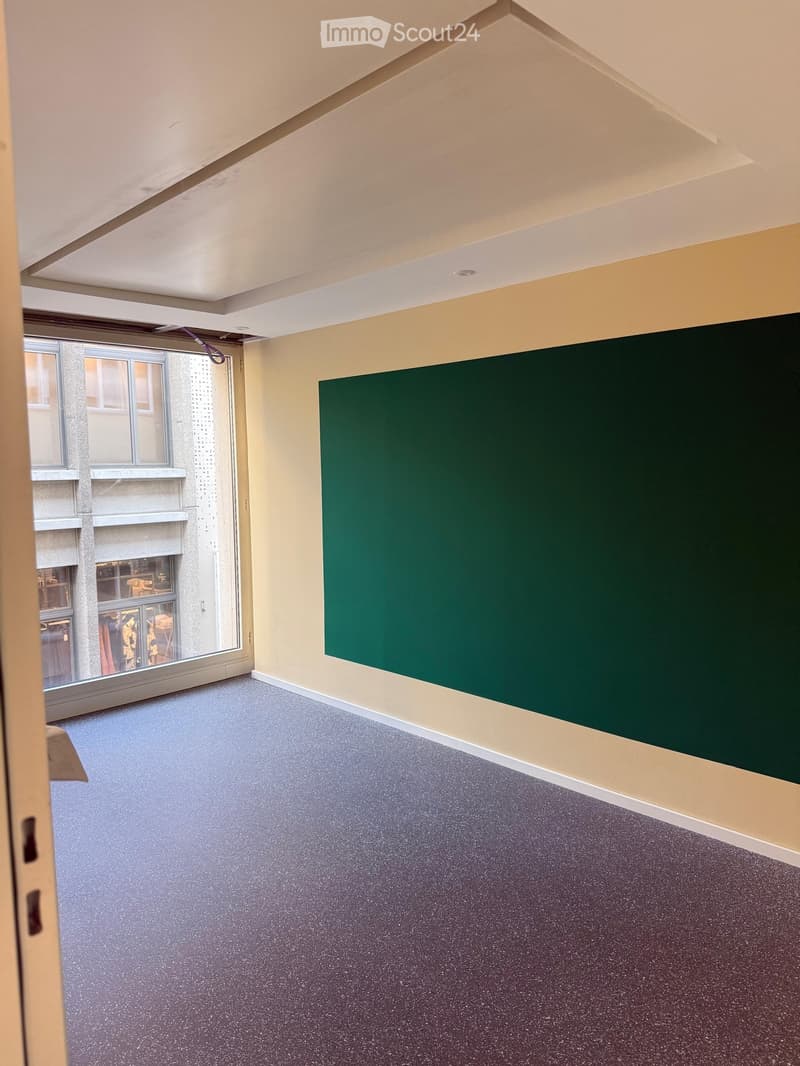 Empty room with light yellow walls, dark grey carpet, large window with view of building exterior, and green chalkboard on one wall.