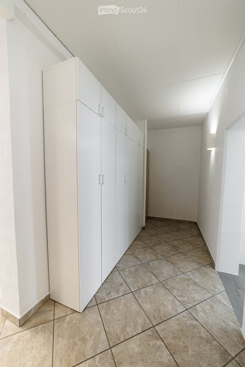 Empty hallway with white walls, beige tiled floor, built-in white storage units with silver handles, and recessed lighting.