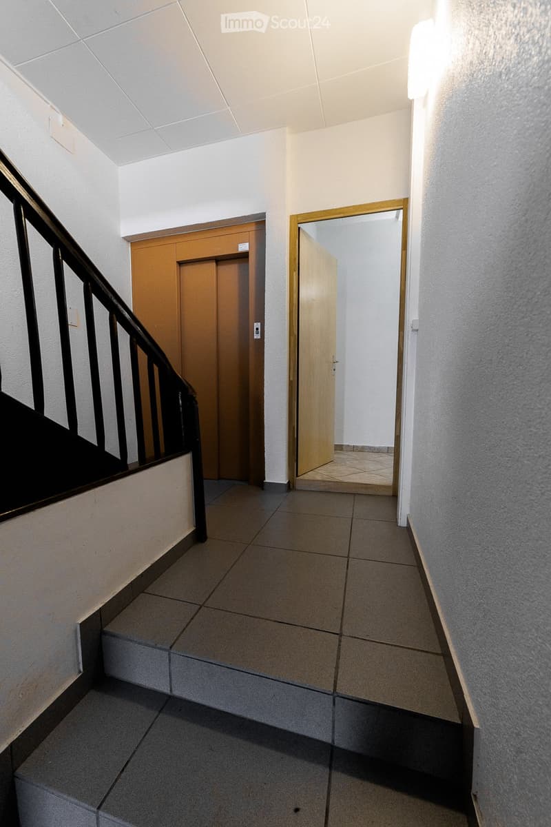 A clean, modern hallway with tiled flooring, white walls, and a staircase with a black railing on the left. A wooden door is visible at the end of the hallway, leading to another room.