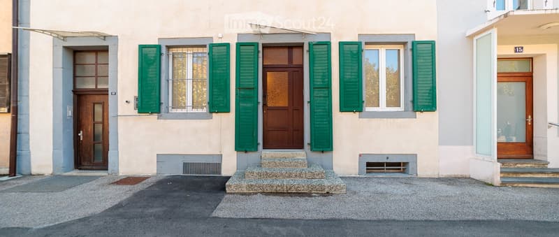 A two-story residential building with beige walls, green shutters, and multiple wooden doors with glass panels. The entrance features a small set of stone steps leading to a central door.
