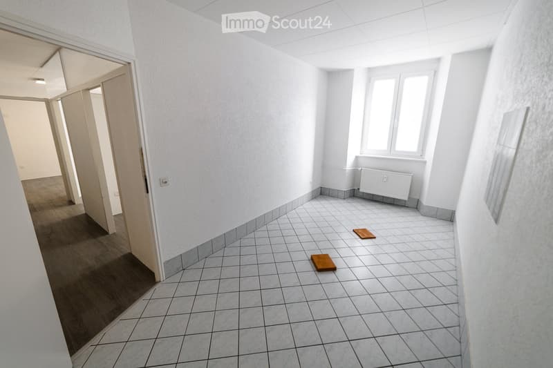 Empty hallway with white walls, tiled floor, and a radiator. Two small wooden boards are placed on the floor.