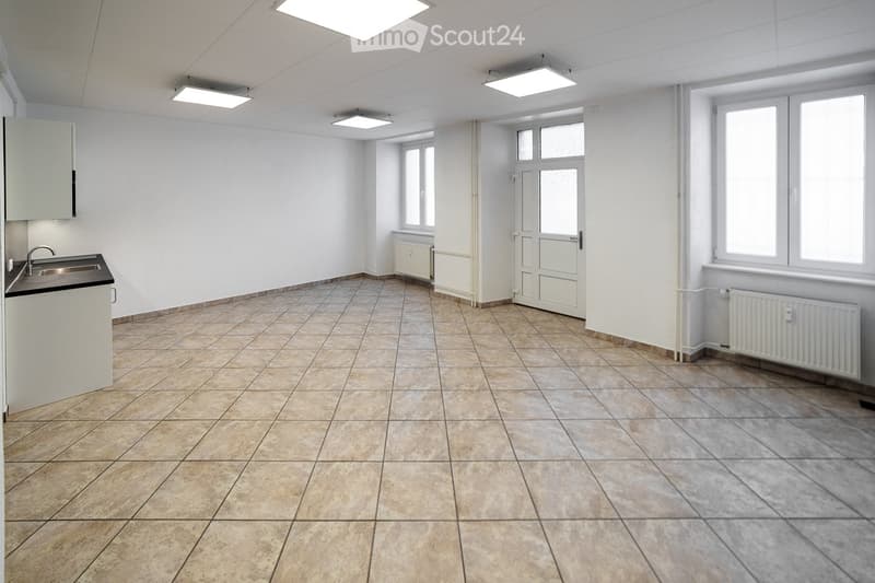 Empty room with tiled floor, white walls, and a small sink area. The room has several windows and a door leading to another space.