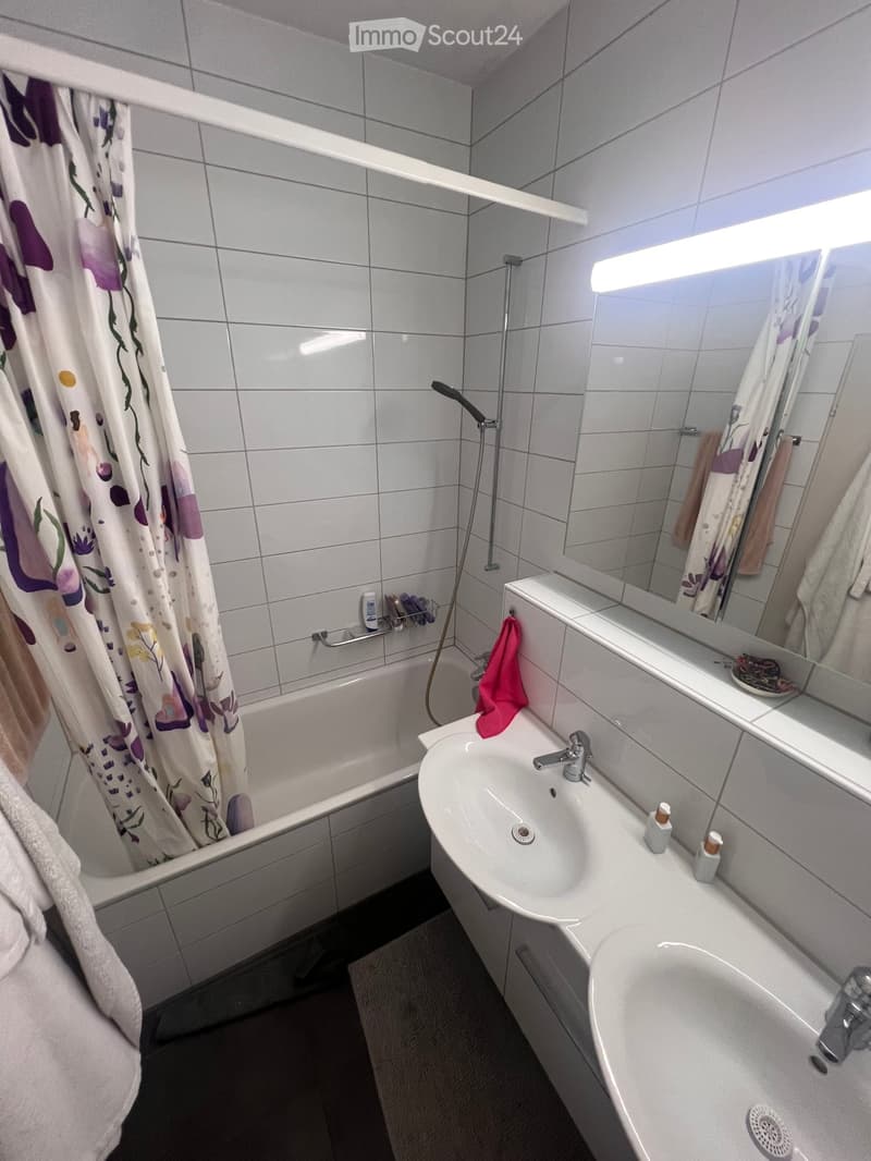 Modern bathroom with a bathtub, double sink, large mirror, and shower area with a curtain. Towels and toiletries are visible.