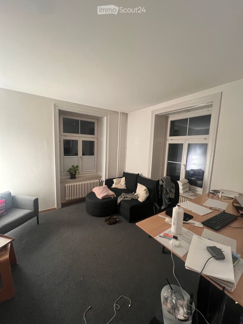 A furnished room with a grey carpet, a black sectional sofa with pillows, a grey armchair, a wooden desk with a laptop, keyboard, and various papers, a small plant on the windowsill, and a radiator below the window.