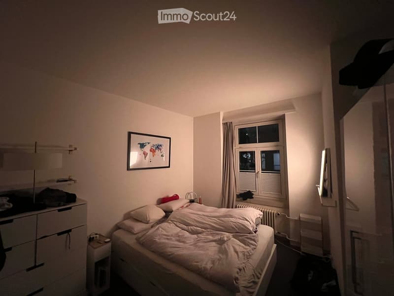 A small, neatly arranged bedroom with a single bed, pillows, a nightstand, a drawer unit, a picture frame on the wall, and a window with curtains.