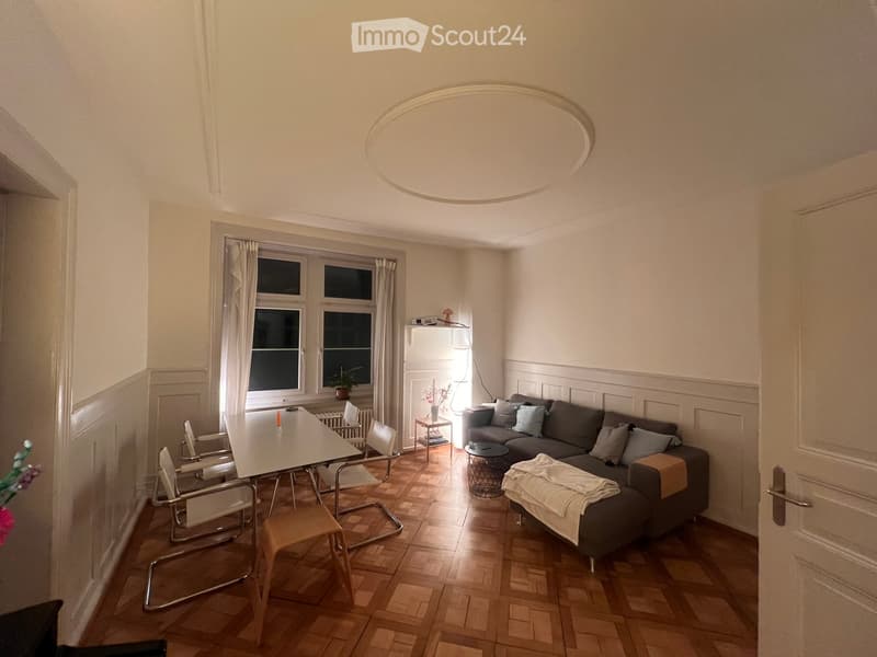 Living room with wooden flooring, large window with curtains, grey sectional sofa, small table, chairs, side table, and floor lamp.