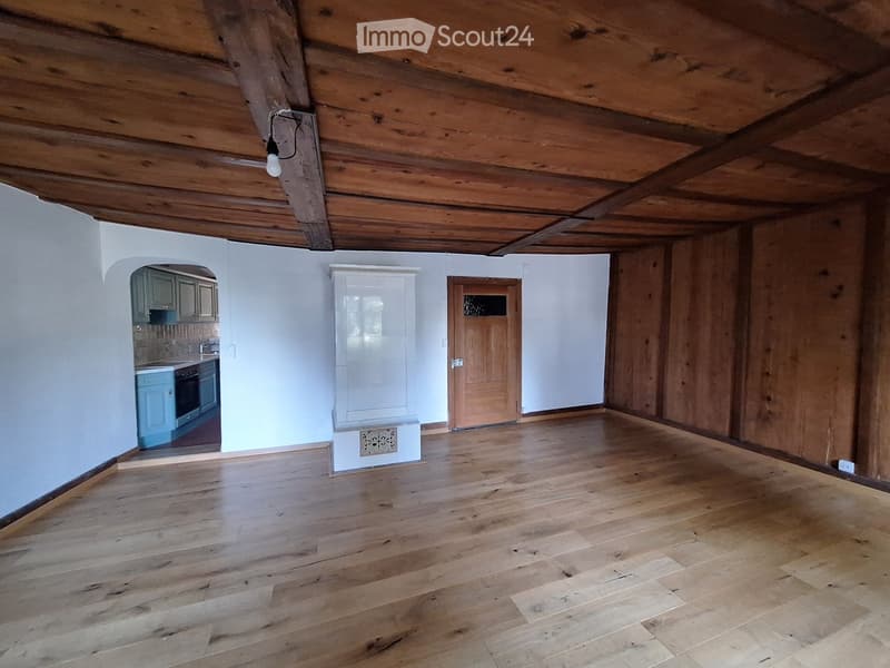 Empty room with wooden floor and exposed wooden beams on the ceiling. There is a doorway leading to another room with cabinets and appliances visible.