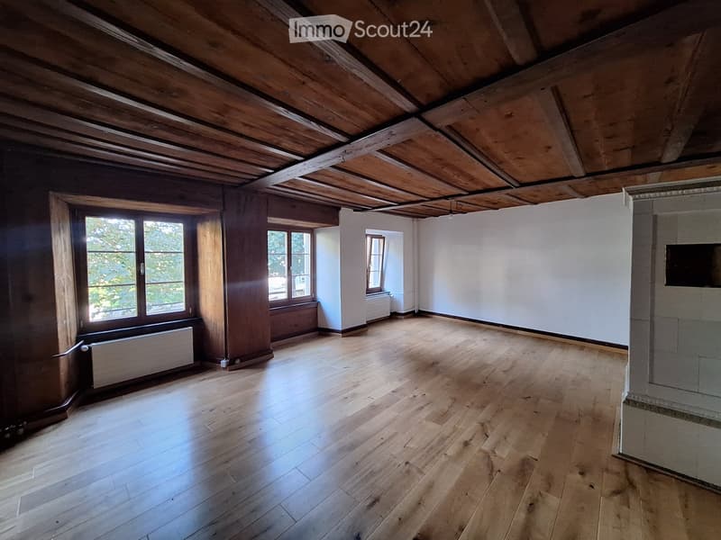 An empty attic room with wooden floors, exposed wooden beams, and large windows allowing natural light to enter.