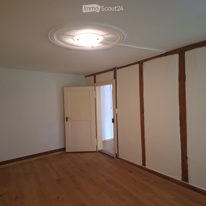 Empty room with wooden flooring, white walls, and a ceiling light fixture. A door leads to another room.