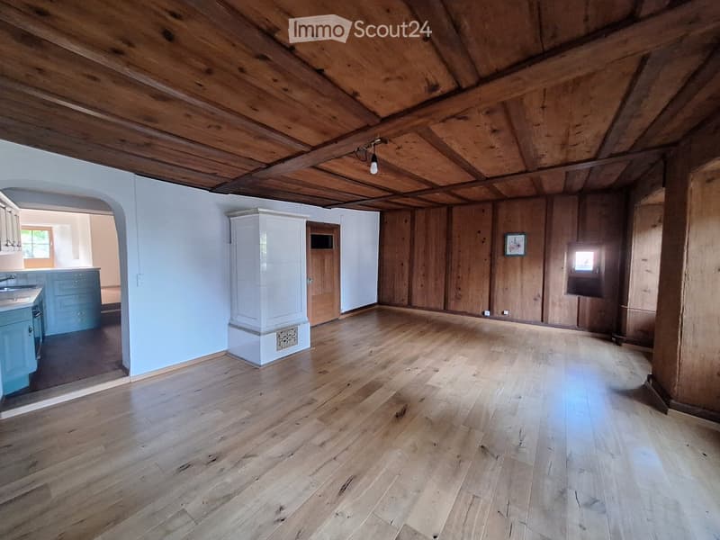 An empty attic space with wooden beams on the ceiling, wooden walls, and a polished wooden floor. The room has exposed wooden beams and a single light fixture hanging from the ceiling. There is a doorway leading to another room with blue cabinets and a si