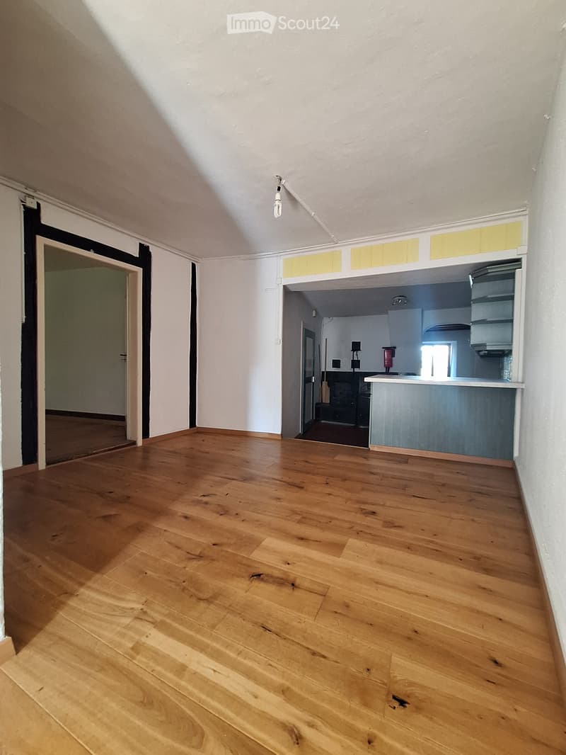 Empty room with wooden flooring, white walls, and a doorway leading to another room. The adjacent room has a kitchen area with cabinets and a countertop.