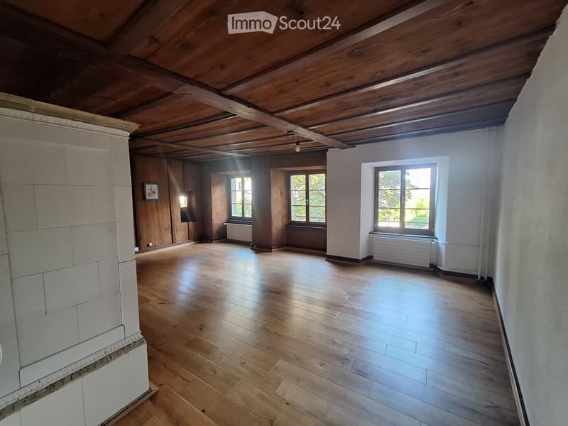Empty room with wooden floors, exposed wooden ceiling beams, and large windows allowing natural light. The walls are painted white, and there are radiators beneath the windows.