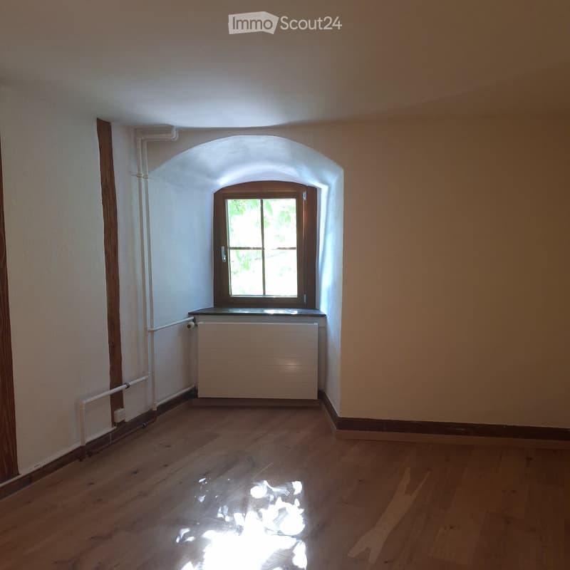 Empty room with wooden flooring, white walls, and a single window with wooden frame. Radiator is visible below the window.