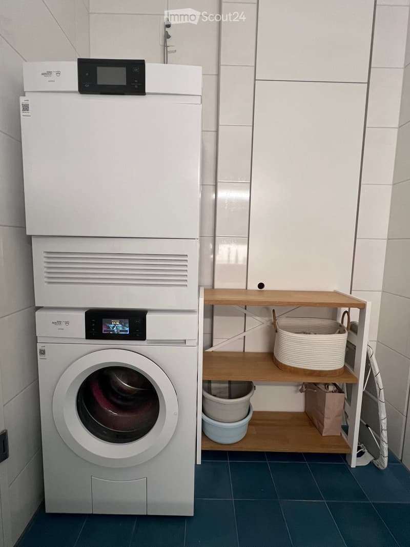 A compact bathroom with a white washing machine and dryer unit, a wooden shelf holding a basket, cup, and handbag, and blue tiled flooring.