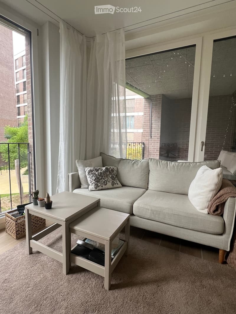 A modern living room with a grey sectional sofa, a light wood coffee table, and a small side table with decorative items. The room features large windows with sheer curtains and a view of a balcony and outdoor area.